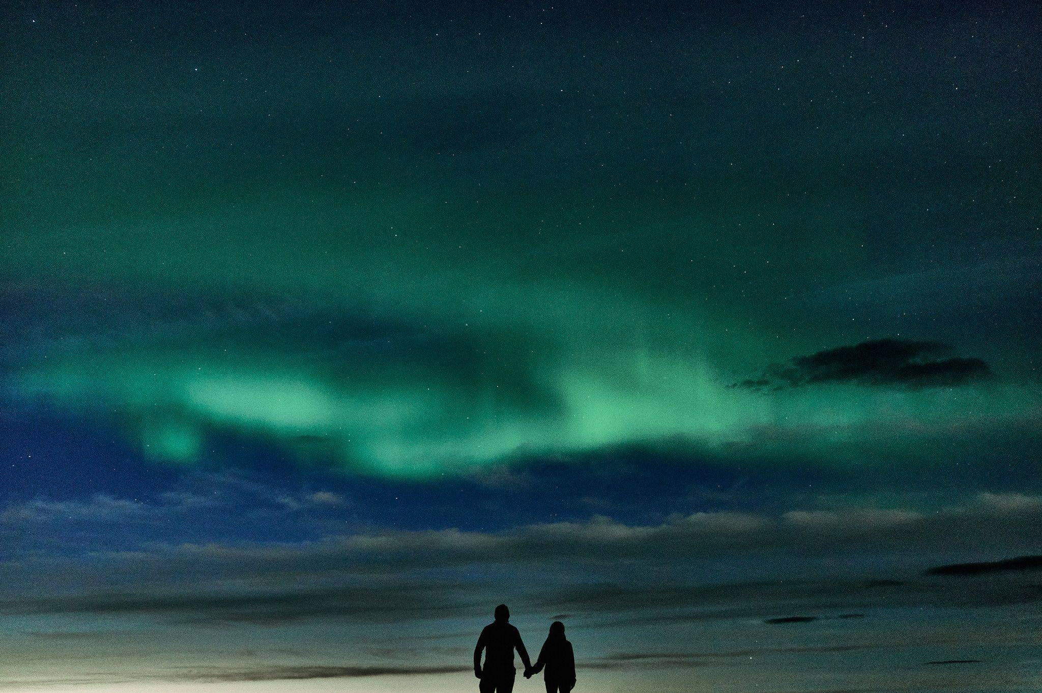Couple holding hands under vibrant green northern lights in iceland’s night sky.